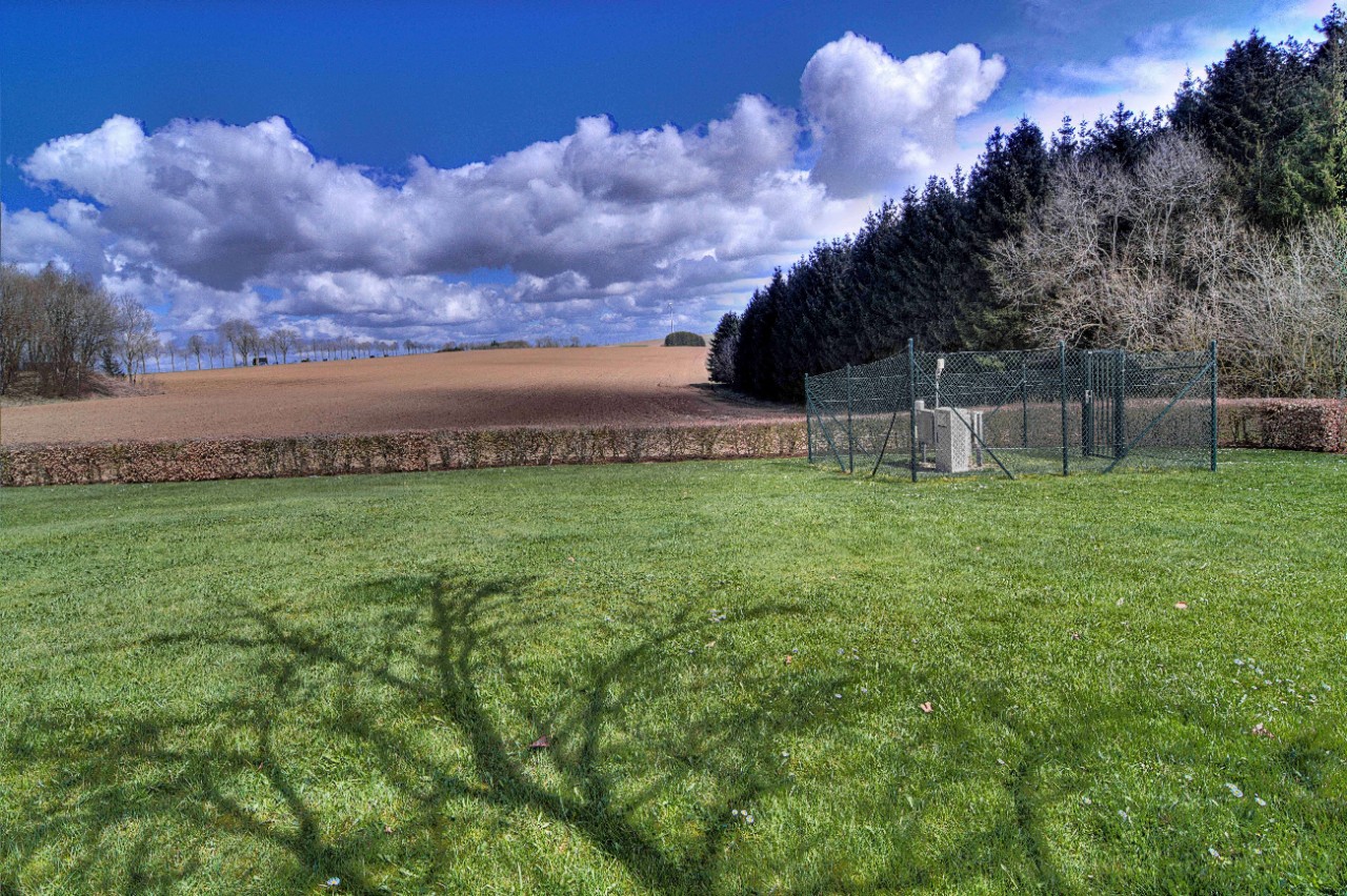 Eine Wetterstation ist auf einer Wiese installiert. Dahinter ist ein Weizenfeld zu sehen, rechts davon ein Tannenwald. Am blauen Himmel ziehen Wolken auf.